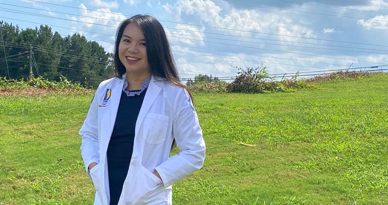 PCOM Georgia pharmacy student Diana Tran (PharmD '21) smiles and wears her white coat outside in a grassy field.