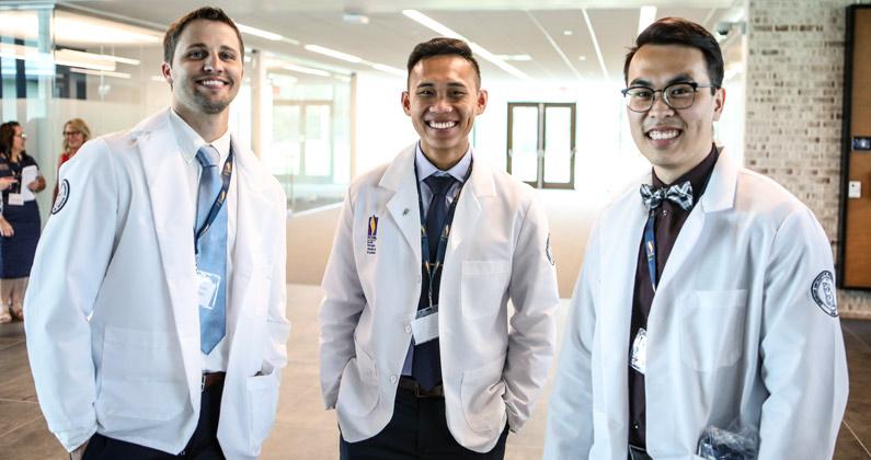 Three PCOM South Georgia students wear their white coats and smile in a campus lobby.