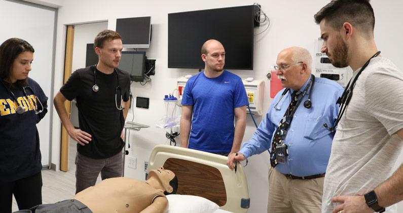 A professor speaks to students as they learn to examine a patient mannequin with their stethoscopes.