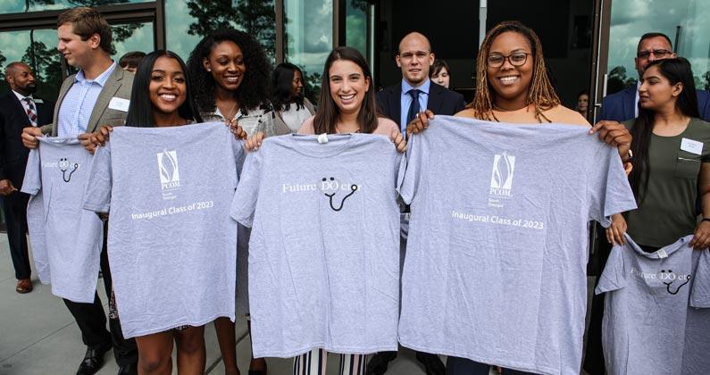 Med students smile and hold up t-shirts outside of PCOM South Georgia's front doors during NOM Week 2020 in April.