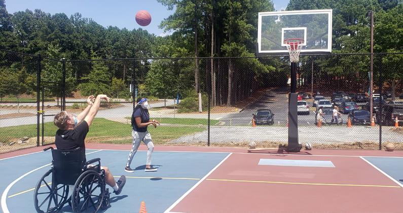 A PCOM Georgia physical therapy student assists a classmate using a wheelchair to shoot a basketball on an outdoor court.