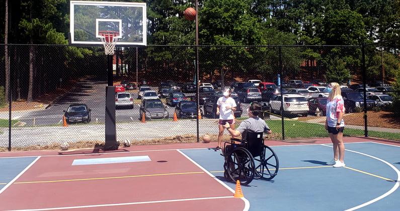 PCOM Georgia's PT students watch as a classmate in a wheelchair shoots a basketball on an outdoor court.
