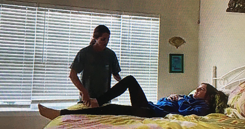 For a virtual class presentation, a physical therapy student demonstrates a stretch on her roommate as she lies down on a bed.