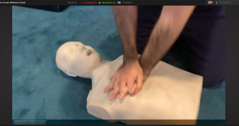 A medical student demonstrates CPR compressions on a mannequin for girl scouts during a virtual learning event.