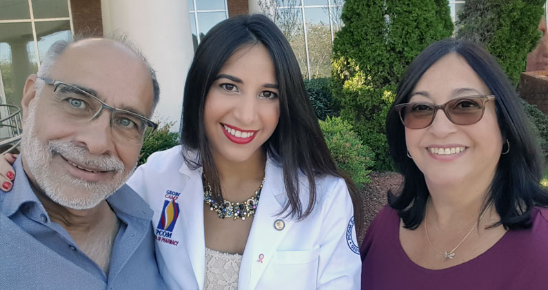 Recent PCOM Georgia graduate Vivian Flores, PharmD, dons her physician white coat and smiles beside her parents.